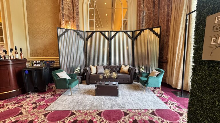 Elegant lounge area featuring a brown tufted leather sofa, green velvet accent chairs, gold décor, and a corrugated metal backdrop on a patterned ballroom carpet.
