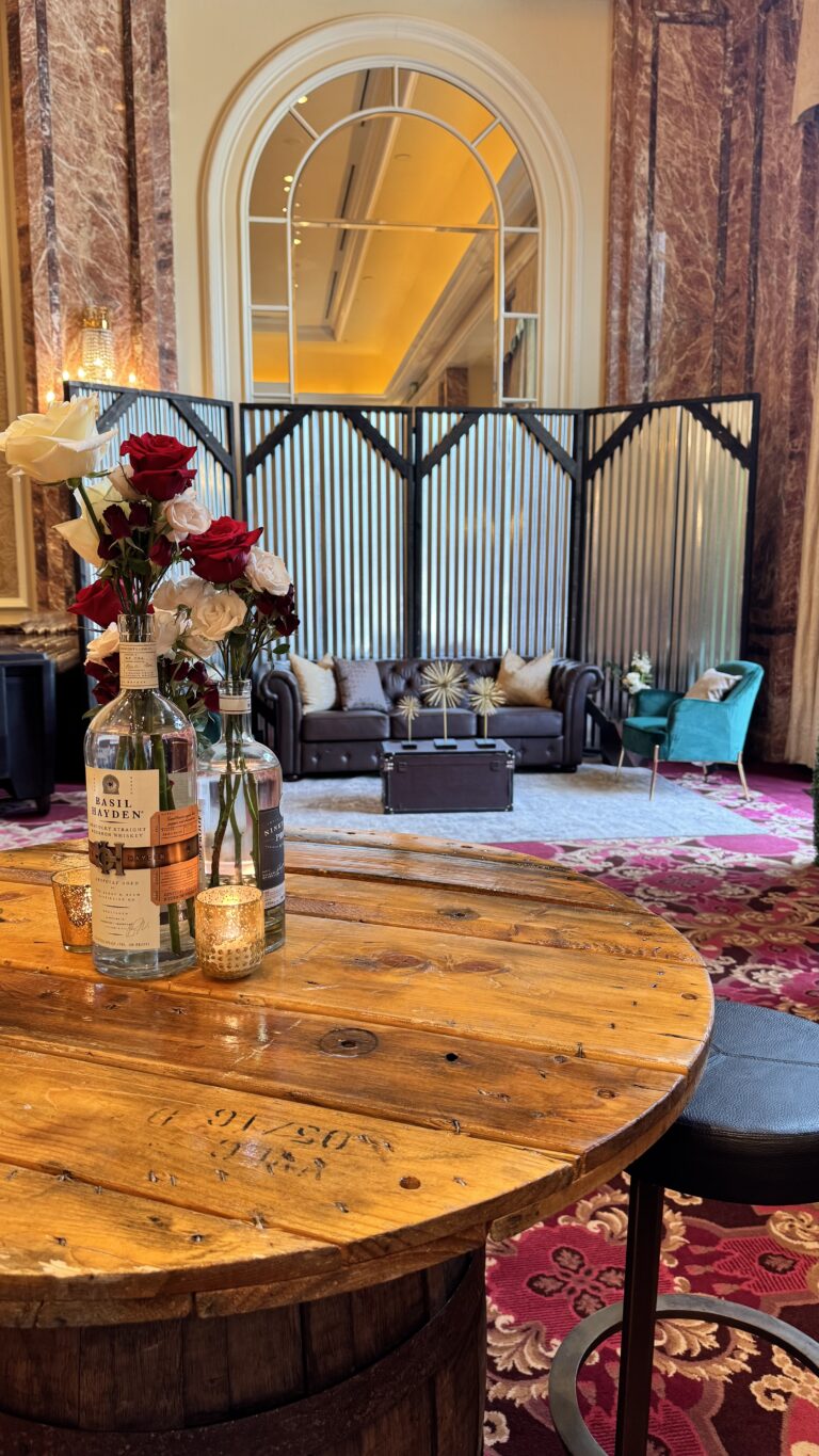 Rustic whiskey bottle vases filled with red and white roses on a wooden table, accented by gold votive candles in an elegant ballroom setting.