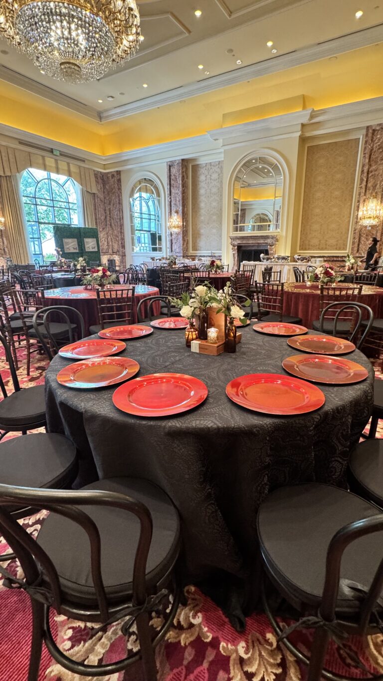 Round dining table with black linen, red charger plates, and floral centerpiece inside a grand ballroom with chandeliers and gold detailing.