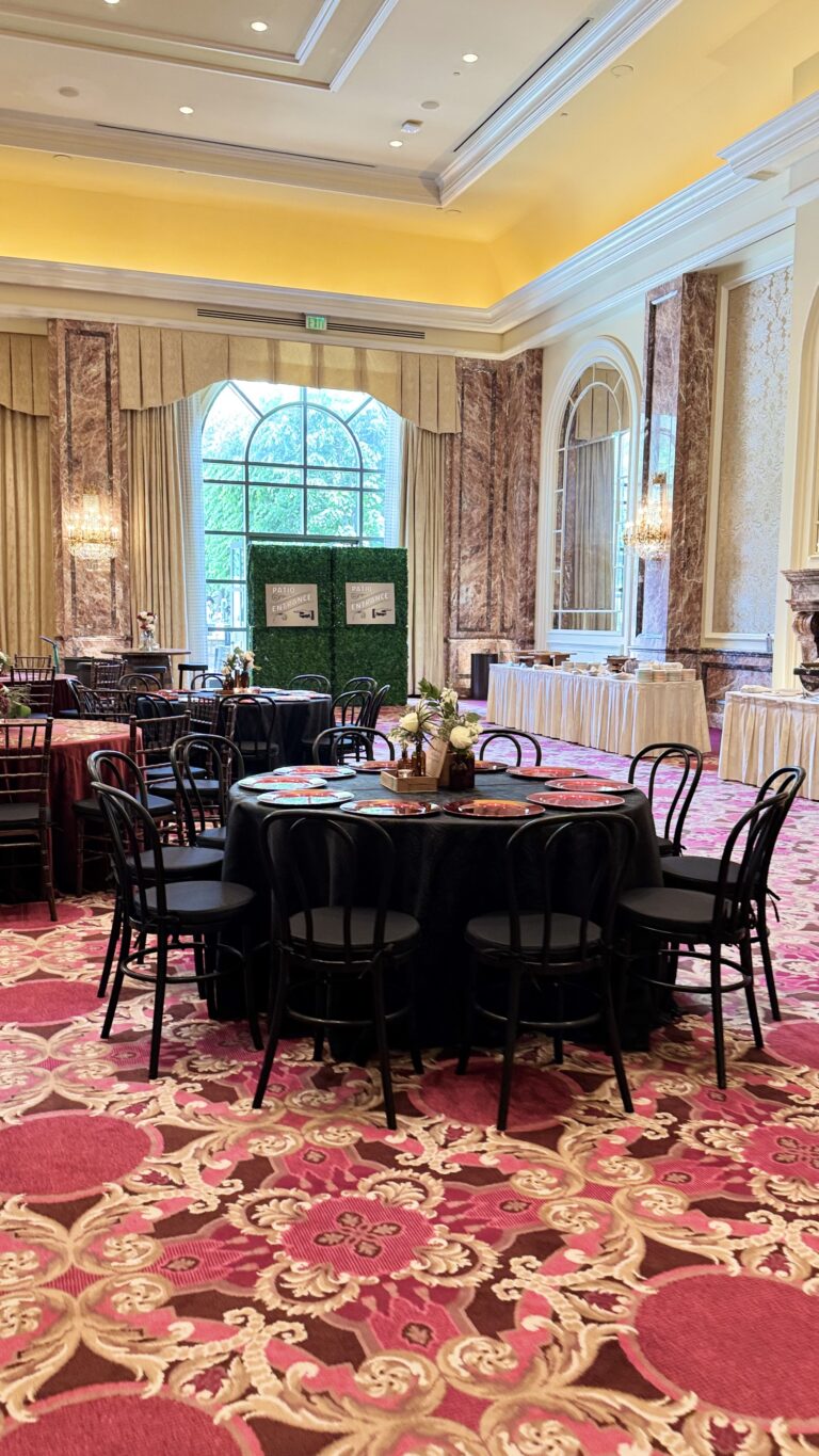 Luxurious ballroom setup featuring black and burgundy linens, floral centerpieces, and gold-accented chargers beneath chandeliers and tall arched windows.