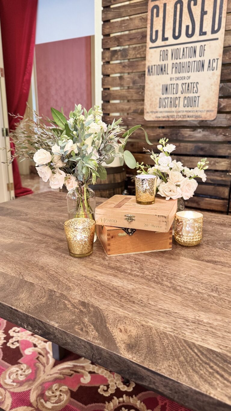 Elegant tabletop display featuring white floral arrangements, gold votive candles, and vintage cigar boxes against a wooden backdrop with a “Closed for Violation of National Prohibition Act” sign.