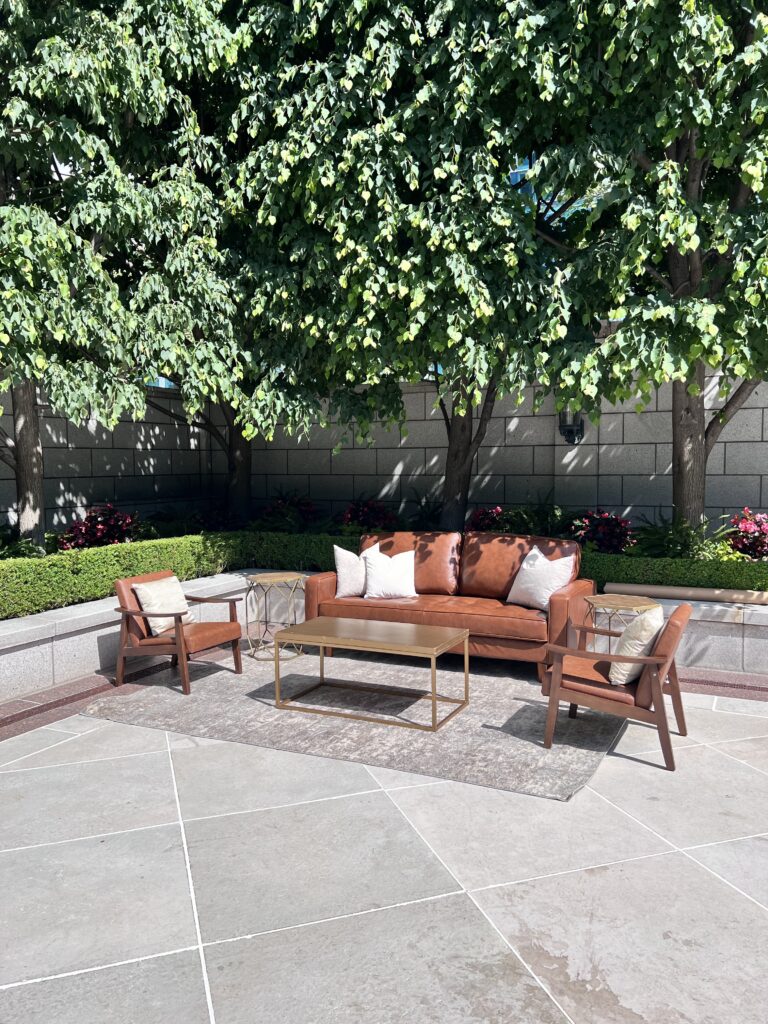 Brown leather sofa and matching armchairs arranged outdoors on a rug under green trees with accent tables and pillows, styled for an elegant event.