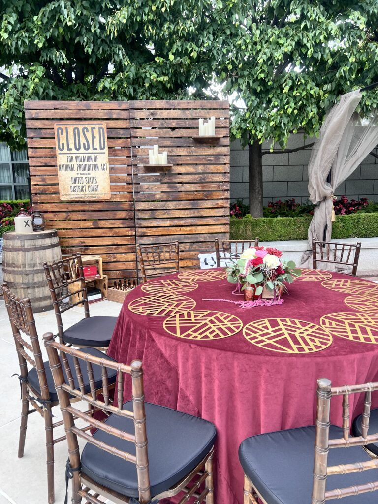 Elegant outdoor event setup featuring a rustic Prohibition-era backdrop, burgundy velvet tablecloth with gold placemats, and floral centerpiece styled by In The Event.