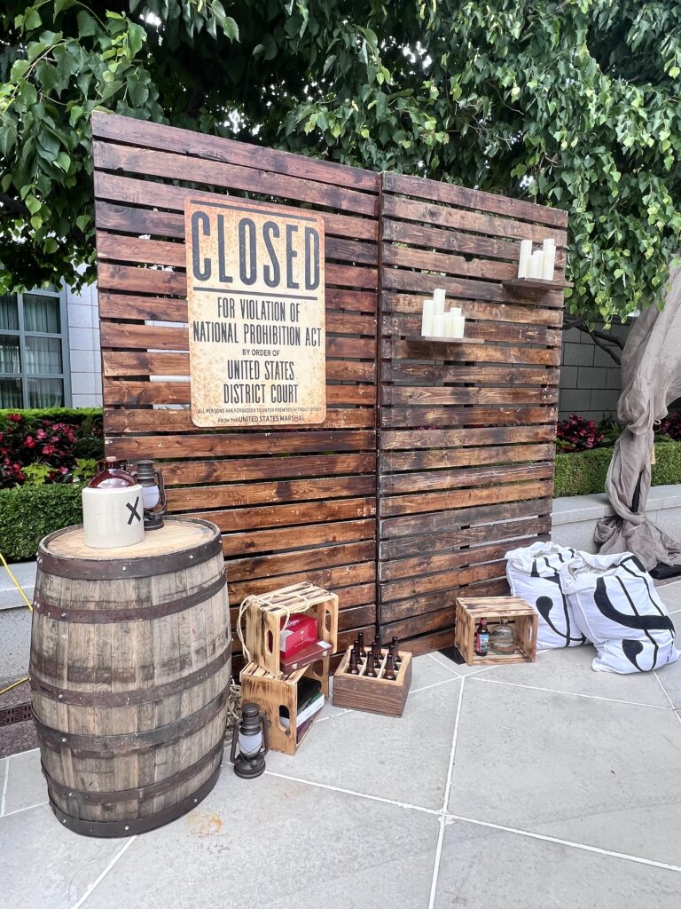 Rustic wooden backdrop with “Closed for Violation of National Prohibition Act” sign, whiskey barrels, lanterns, and vintage props styled by In The Event.