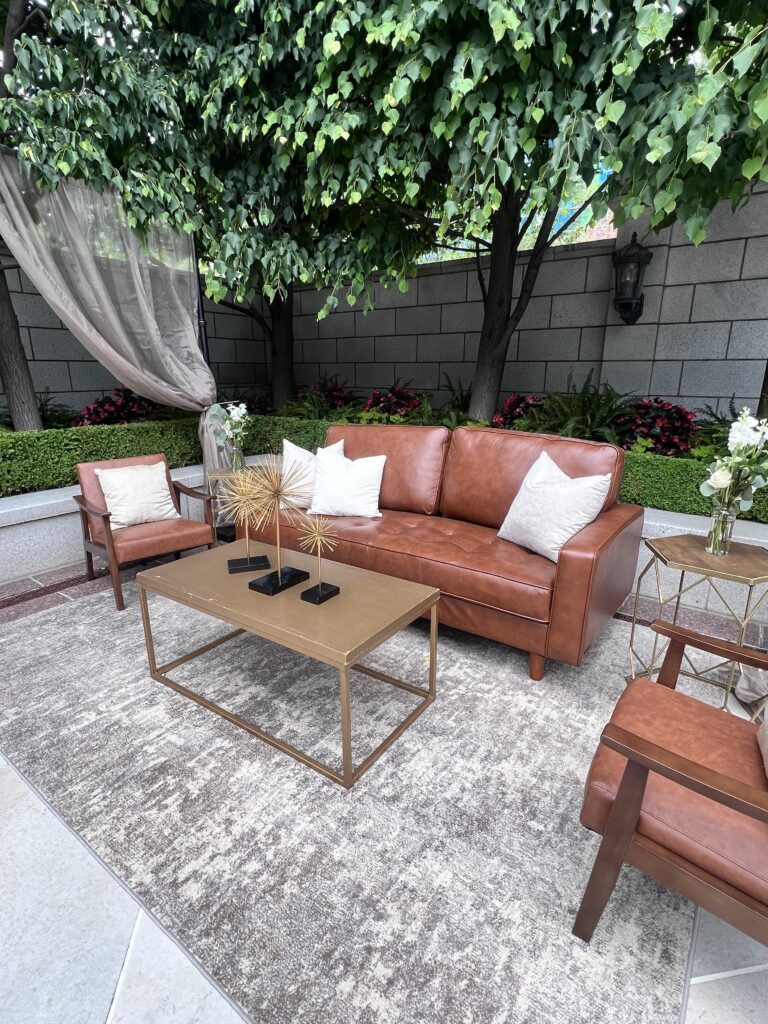 Stylish outdoor lounge featuring cognac leather sofa and chairs, modern coffee table, and soft neutral rug under tree canopy styled by In The Event.