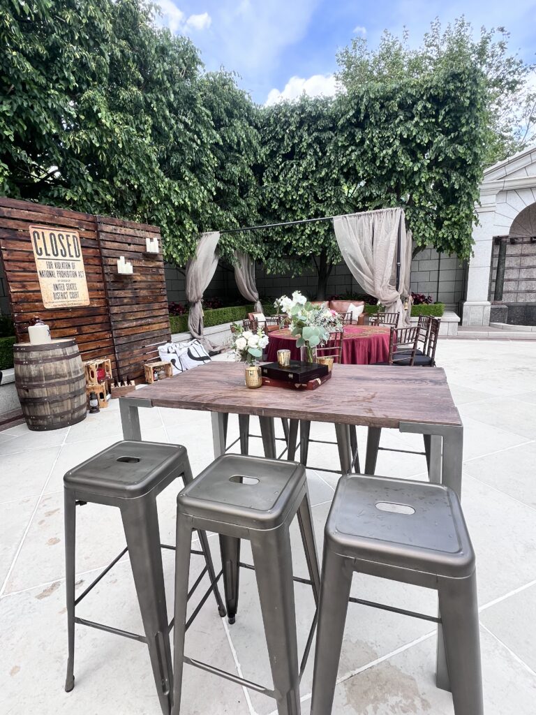 Outdoor event scene featuring a “Closed for Violation of National Prohibition Act” backdrop, metal barstools, wood tables, and burgundy lounge drapery styled by In The Event.