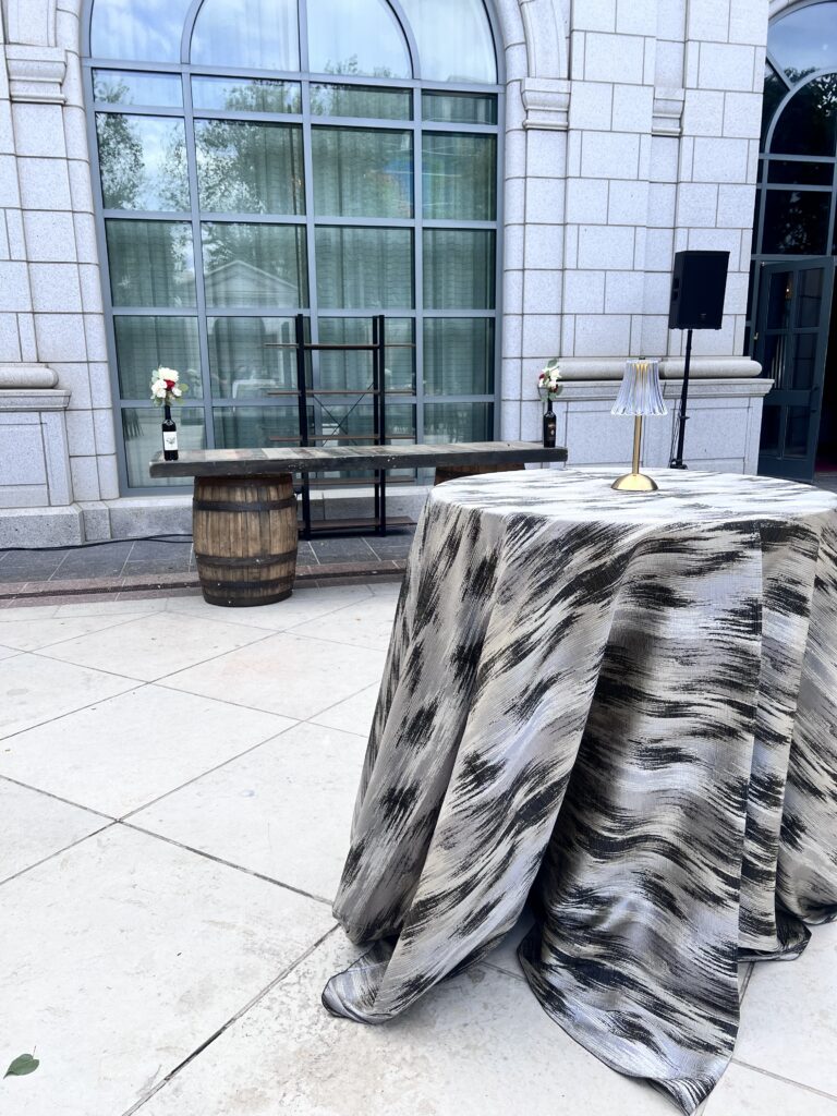 Elegant outdoor cocktail setup featuring a high-top table with black and silver patterned linens, whiskey barrel bar, and floral accents styled by In The Event.