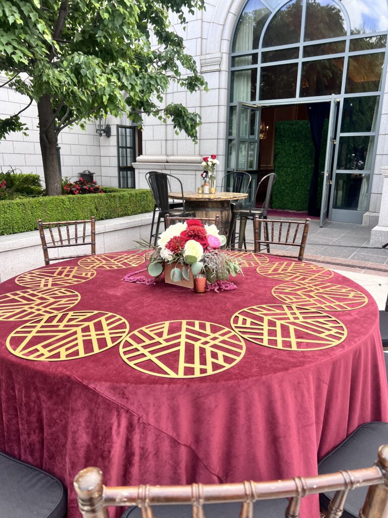Elegant outdoor event setup featuring a burgundy velvet tablecloth, gold geometric placemats, and floral centerpiece with roses and dahlias styled by In The Event.