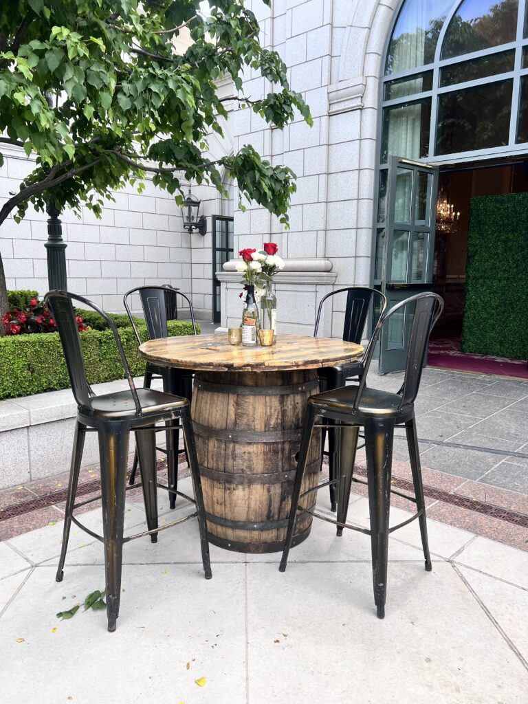 Rustic whiskey barrel cocktail table with metal barstools and rose centerpiece setup outside a grand venue entrance, styled by In The Event.