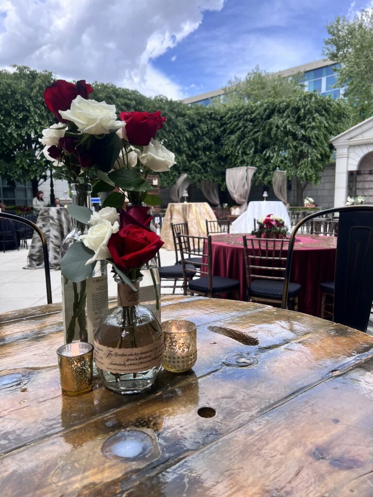 Close-up of red and white rose arrangement in glass bottles with gold votive candles on a rustic whiskey barrel table during an outdoor event by In The Event.