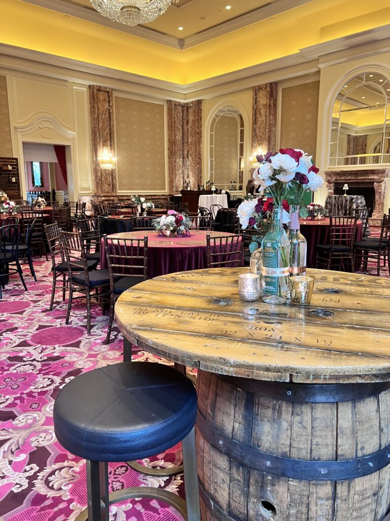 Rustic whiskey barrel cocktail table with floral centerpiece and votive candles in an elegant ballroom with chandeliers and burgundy linens styled by In The Event.