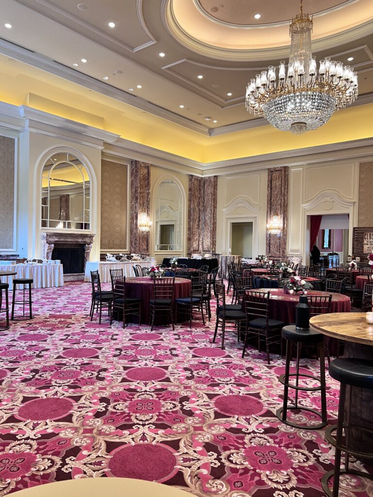 Lavish ballroom with crystal chandeliers, patterned carpet, and round tables with rich burgundy and black linens styled by In The Event.