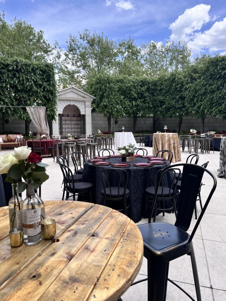 Stylish outdoor event setup featuring a rustic wood cocktail table with floral decor, round dinner tables with red chargers, and greenery-lined courtyard by In The Event.
