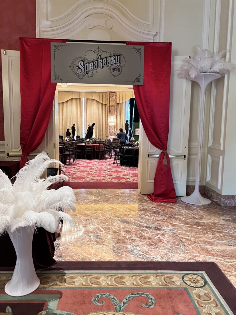 Glamorous event entrance with red drapery, art deco “Speakeasy” sign, and tall white feather decor leading into a vintage-inspired ballroom designed by In The Event.