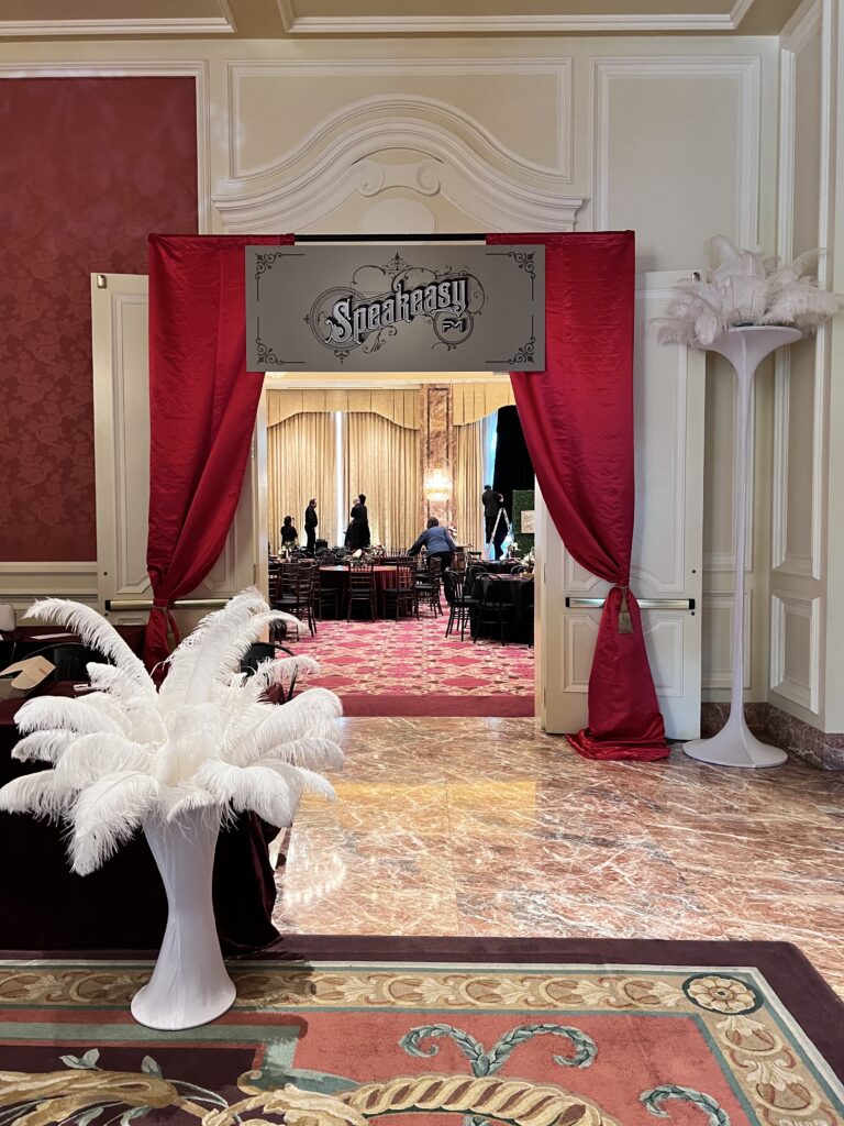 Glamorous event entrance with red drapery, art deco “Speakeasy” sign, and tall white feather decor leading into a vintage-inspired ballroom designed by In The Event.