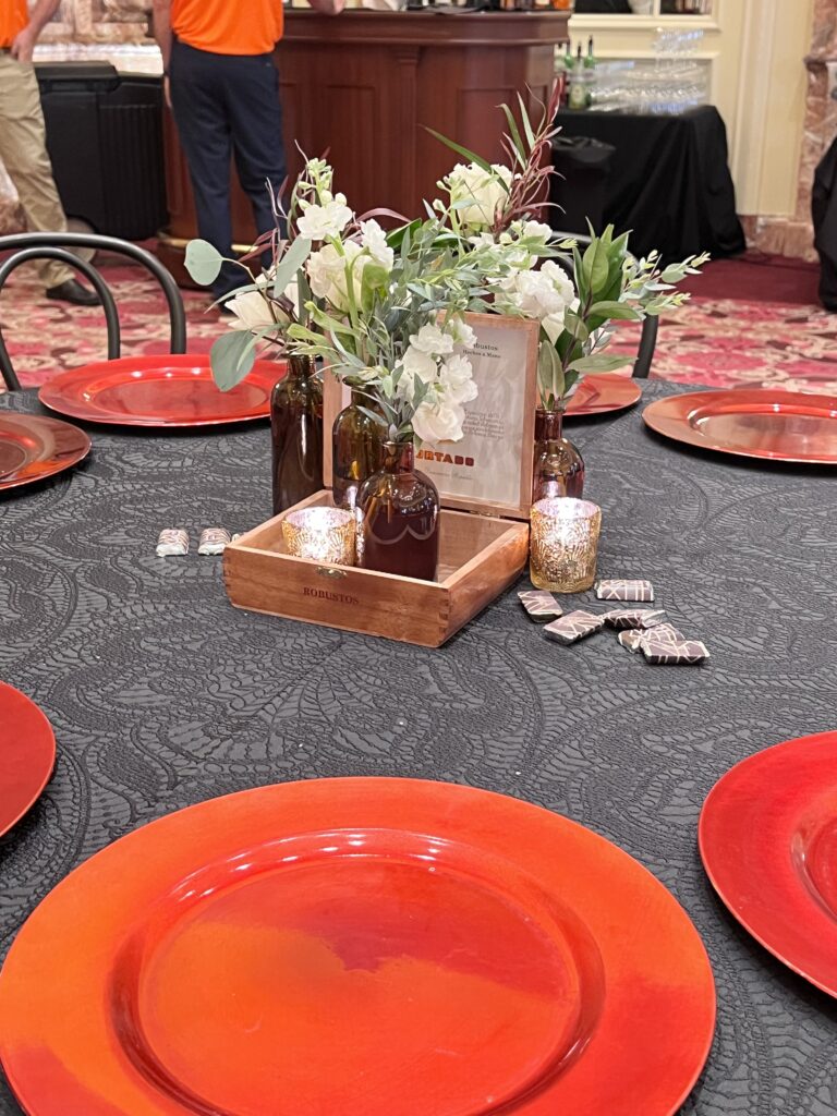 Table centerpiece featuring amber glass vases with white florals, gold votive candles, and a wooden cigar box on a black lace-patterned tablecloth with red charger plates styled by In The Event.