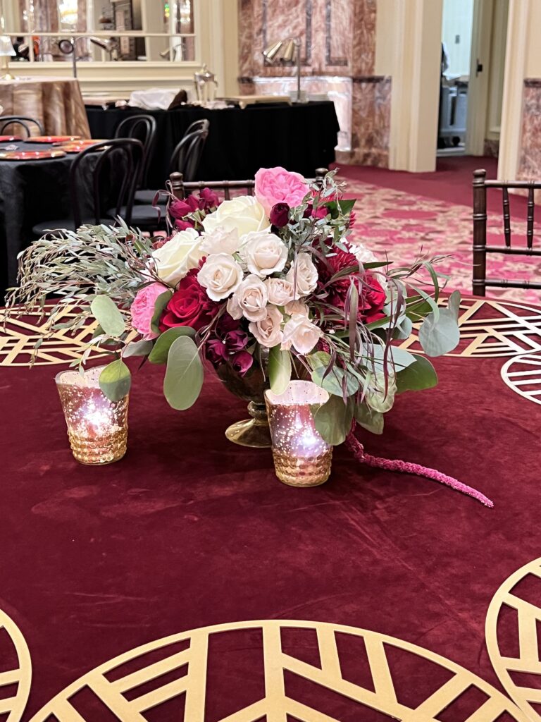 Elegant centerpiece featuring red and white roses with greenery in a gold vase, paired with votive candles on a burgundy velvet tablecloth designed by In The Event.