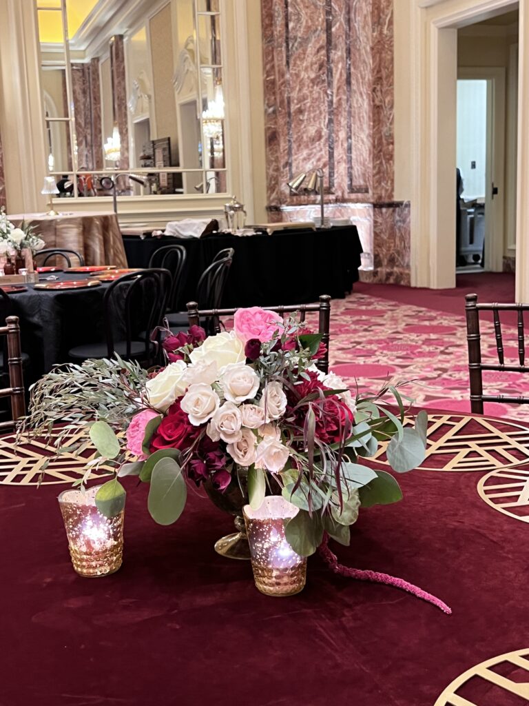 Elegant centerpiece featuring red and white roses with greenery in a gold vase, paired with votive candles on a burgundy velvet tablecloth designed by In The Event.