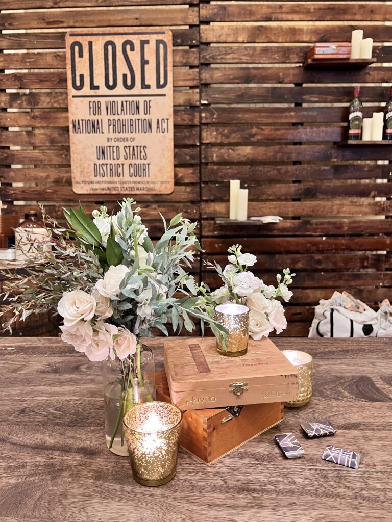 Rustic wood backdrop with “Closed for Violation of National Prohibition Act” sign, whiskey barrels, candles, and floral table arrangement designed by In The Event.
