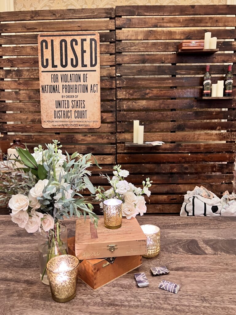Rustic wood backdrop with “Closed for Violation of National Prohibition Act” sign, whiskey barrels, candles, and floral table arrangement designed by In The Event.