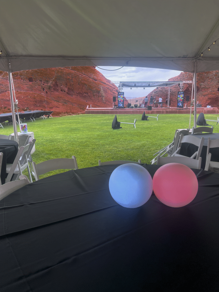 Under-tent table with glowing LED orb centerpieces overlooking stage setup among red rock cliffs at MACU Fest