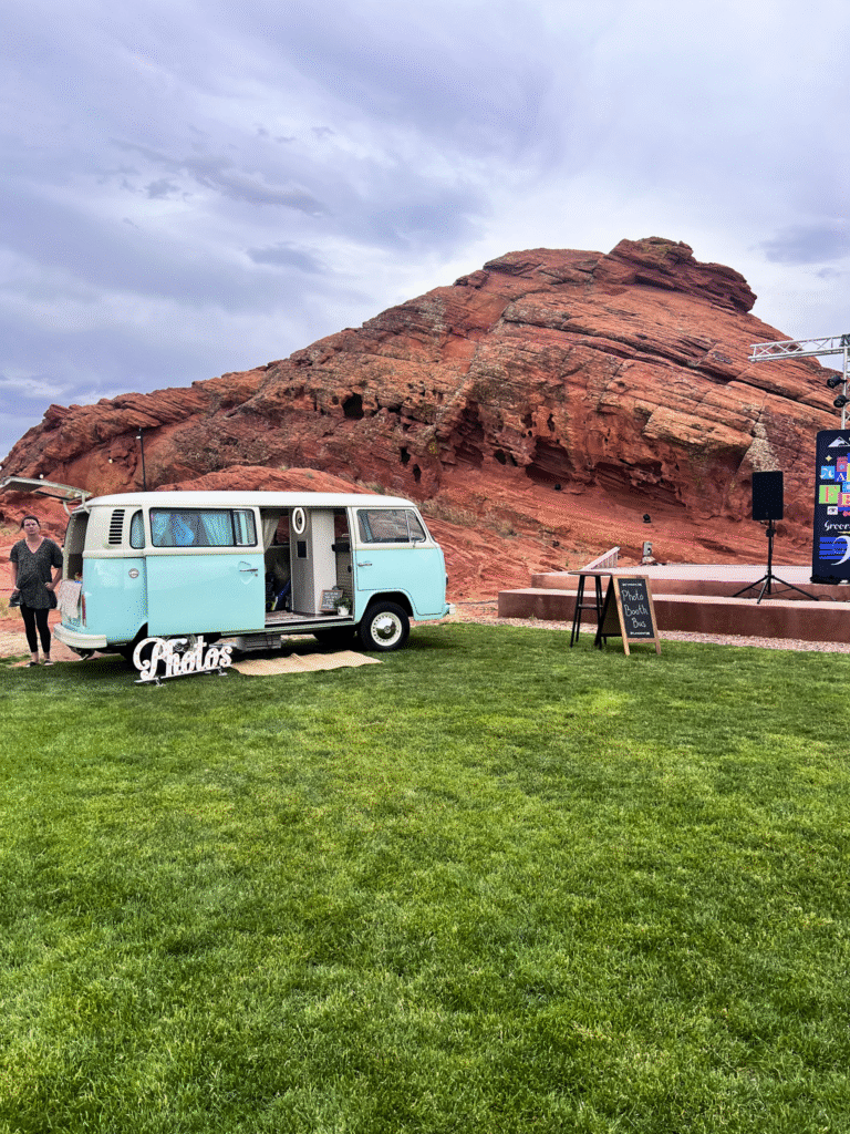 A light blue vintage Volkswagen bus converted into a photo booth, parked on a green lawn with a large red rock formation in the background under a cloudy sky. A "Photos" sign is in front of the bus, and an easel sign for "Photo Booth" is nearby.