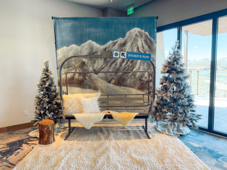 Ski lift-inspired photo backdrop with snowy mountain scene, frosted pine trees, faux fur pillows, and cozy rug creating a winter lodge atmosphere.