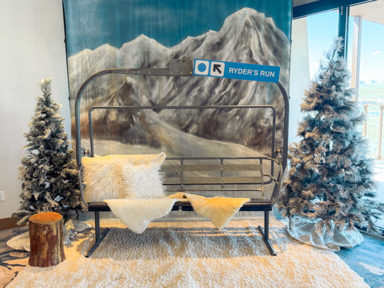 Ski lift-inspired photo backdrop with snowy mountain scene, frosted pine trees, faux fur pillows, and cozy rug creating a winter lodge atmosphere.
