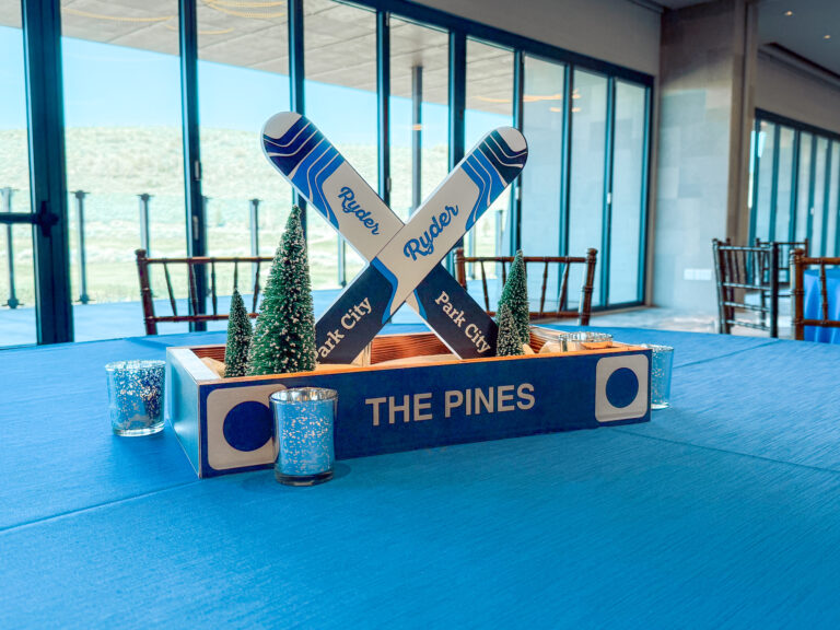 Ski-themed table centerpiece with mini pine trees and custom “Ryder” skis reading “Park City” displayed on a blue tablecloth at an event venue with large windows