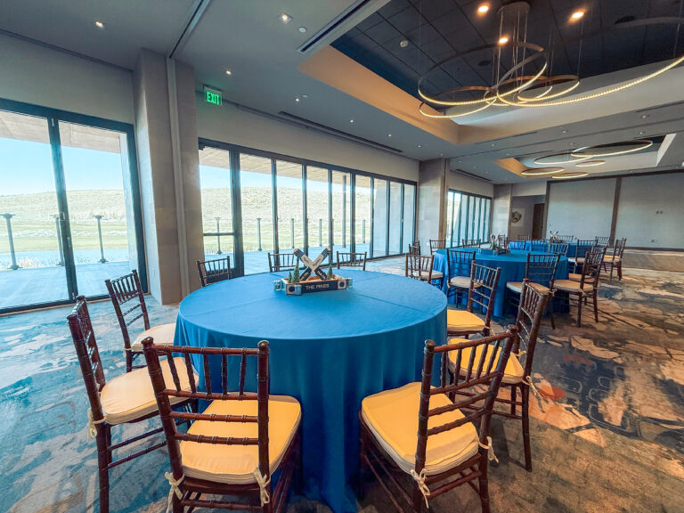 Round event tables with blue linens, wooden chairs, and ski-themed centerpieces in a modern venue with large windows overlooking scenic mountain views.