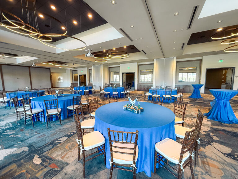 Elegant event space featuring round tables with blue linens, wooden chairs, and custom ski-themed centerpieces under modern ring lighting.