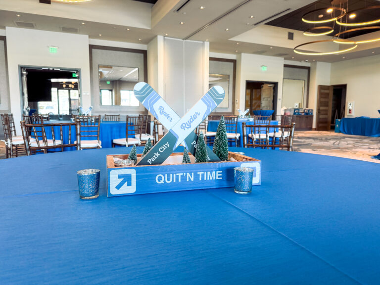 Blue event table featuring a ski-themed centerpiece with crossed skis labeled “Ryder” and “Park City,” mini pine trees, and the phrase “Quit’n Time.”