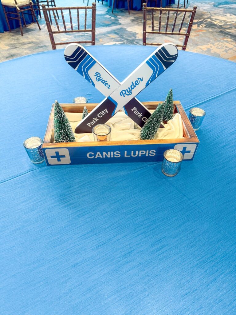 ki-themed table centerpiece with mini pine trees and custom “Ryder” skis reading “Park City” displayed on a blue tablecloth at an event venue with large windows
