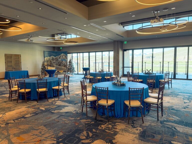 Spacious event venue with blue table linens, wooden chairs, ski-themed centerpieces, and a winter mountain photo backdrop with large windows overlooking scenic views.