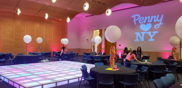 Modern event setup with illuminated LED dance floor, black table linens, colorful balloon centerpieces, and pink uplighting beneath a “Penny ❤️ NY” projection.