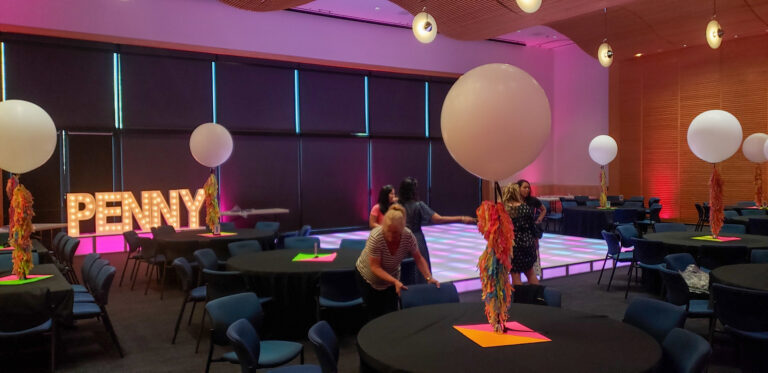 Event setup featuring glowing “PENNY” marquee letters, LED dance floor, and large balloon centerpieces with colorful tassels under pink accent lighting.
