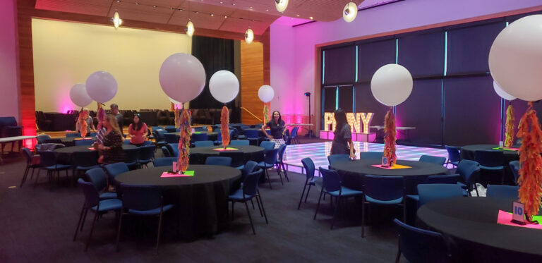 Modern event space featuring black table linens, oversized white balloon centerpieces with colorful tassels, and an illuminated LED dance floor with marquee “PENNY” letters.