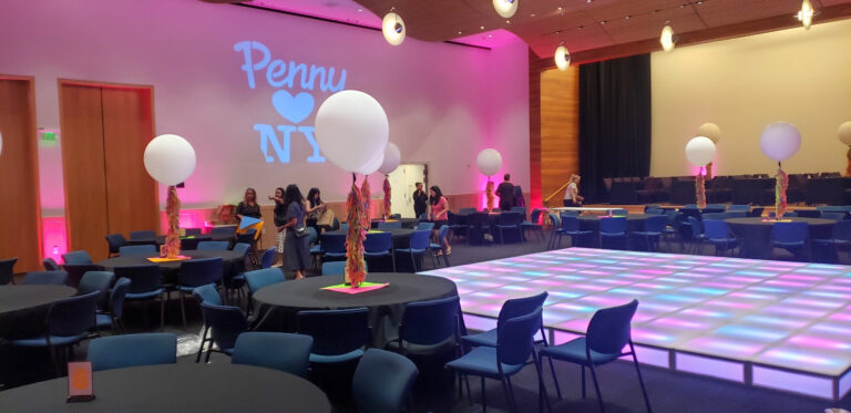 Vibrant event setup with an illuminated LED dance floor, black table linens, neon accents, large balloon centerpieces, and pink uplighting with “Penny ❤️ NY” projected on the wall.