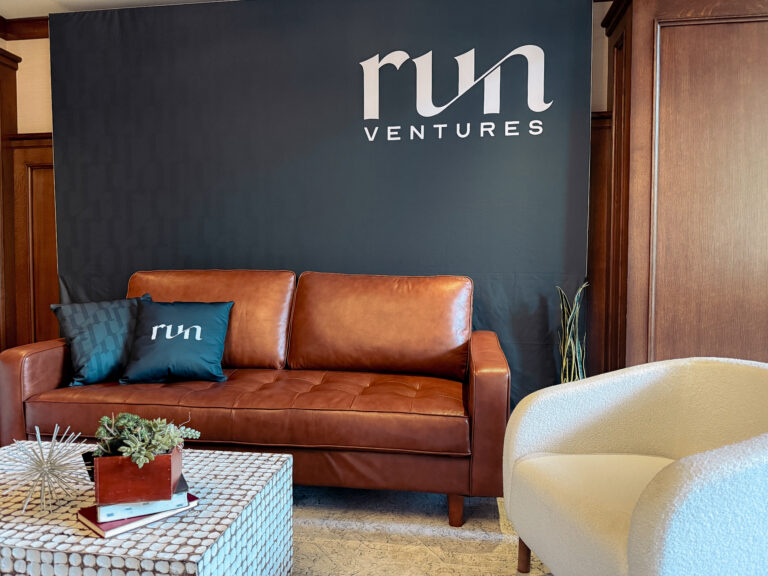 Close-up of event lounge furniture with a brown leather couch, custom branded pillows, and a custom event backdrop featuring the 'run ventures' logo.