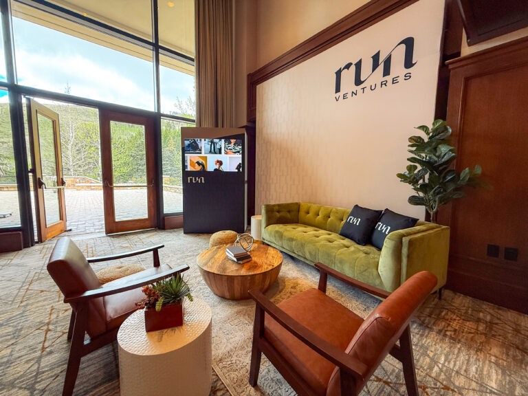 Elegant event lounge furniture with a green velvet sofa, leather armchairs, a round wooden coffee table, custom event signage backdrop, and vertical video rentals screen.