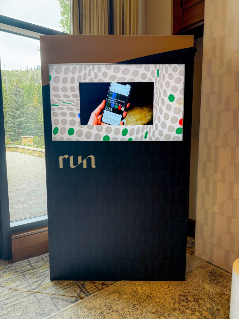 Vertical interactive trade show display kiosk with custom branding and a screen showing a mobile device, set up near a window.