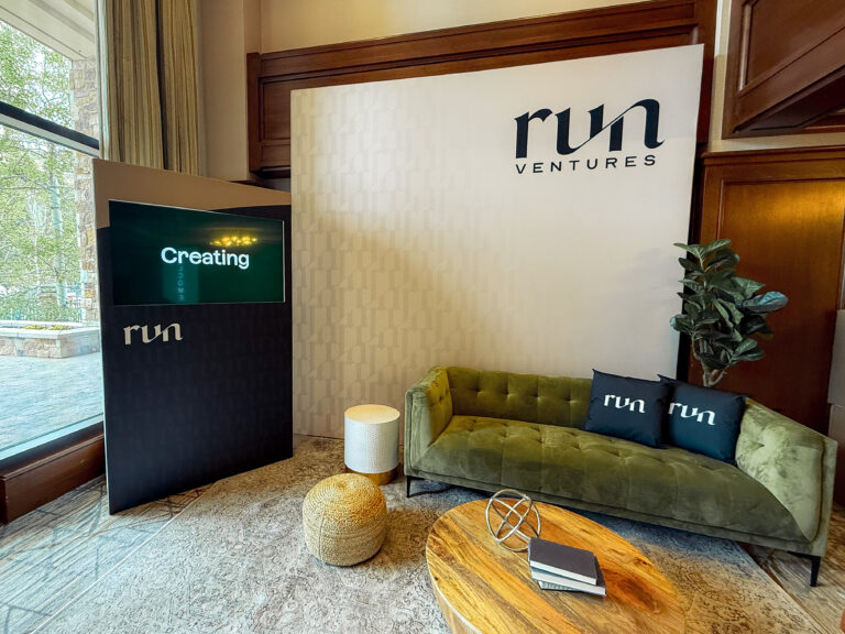 Stylish green velvet sofa in a branded lounge area with custom event backdrops, a vertical video rentals screen, and bohemian table decor.