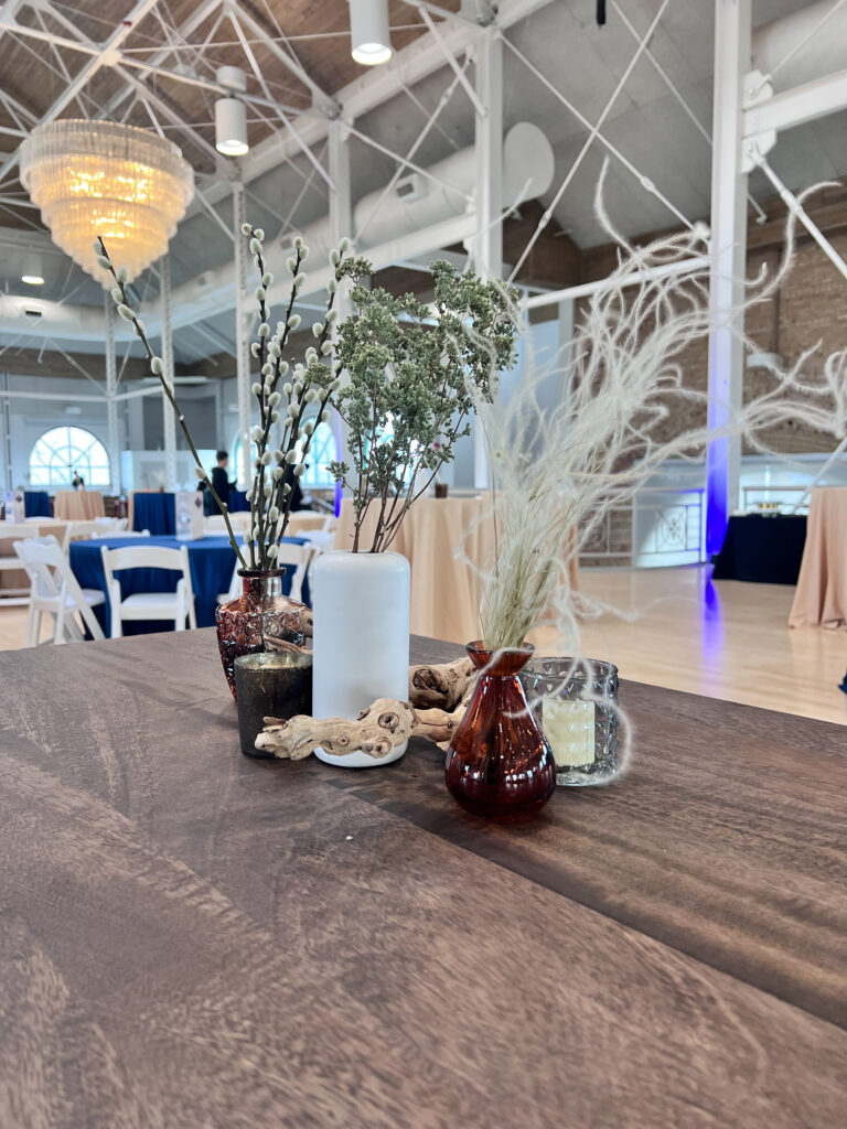 Close-up of dried floral and greenery arrangements in assorted glass vases with candles and driftwood accents on a rustic wooden table styled by In The Event.