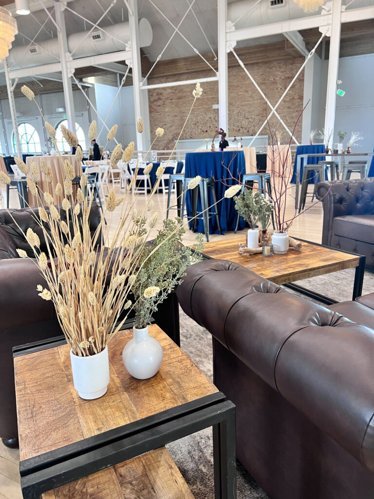 Elegant event lounge featuring brown Chesterfield sofas, rustic wood tables, and minimalist vases with dried floral arrangements styled by In The Event.