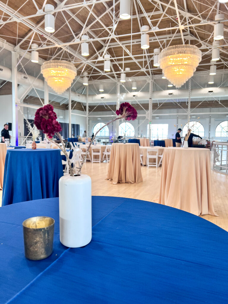 Close-up of a modern table setting with blue linen, white vase floral arrangement, and gold candle votive inside a chandelier-lit venue styled by In The Event.