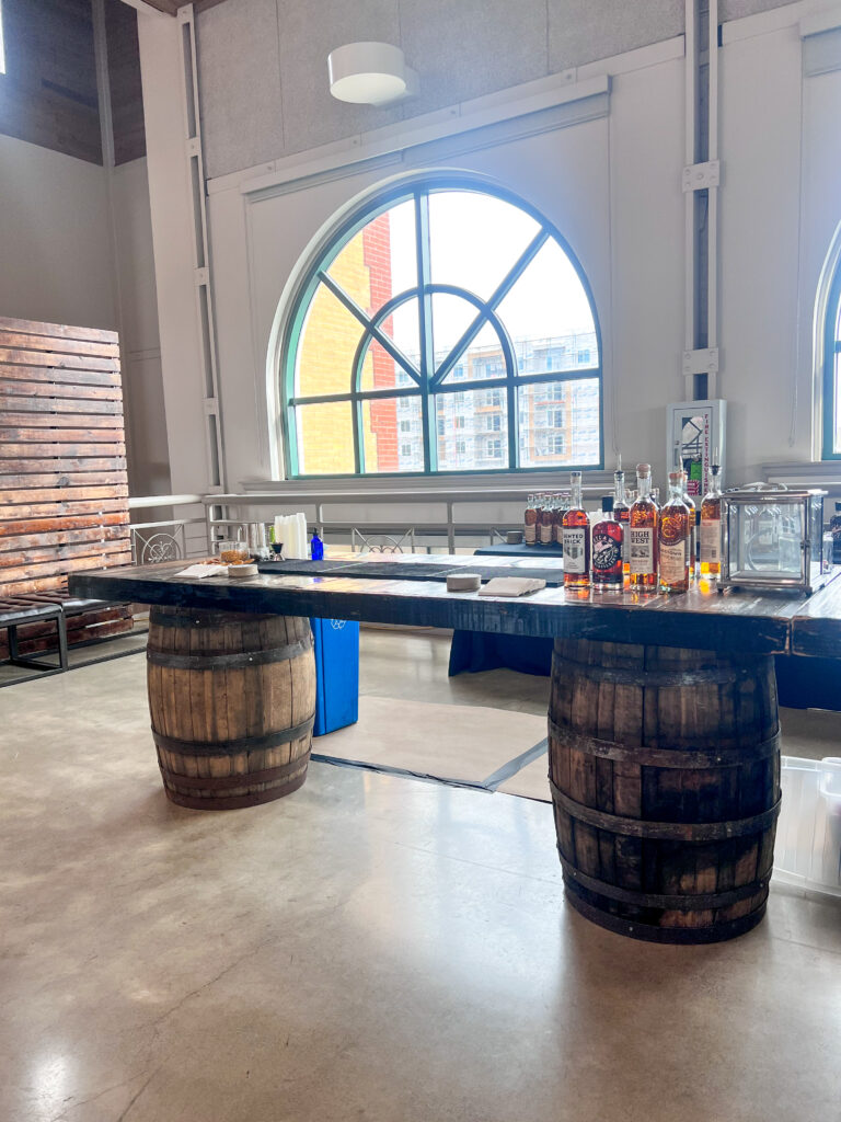 Rustic event bar built from whiskey barrels and dark wood tabletop, featuring assorted liquor bottles and glassware beneath arched windows, designed by In The Event.