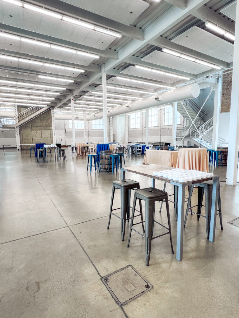 Spacious modern event hall featuring metal barstools, whiskey barrel cocktail tables, and blue and tan linens arranged by In The Event.