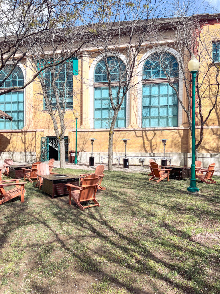 Wooden Adirondack chairs arranged around square fire pit tables with patio heaters outside a historic brick venue, designed by In The Event.