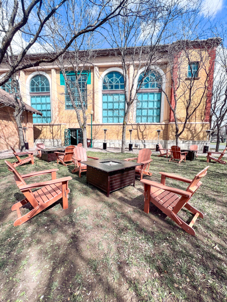 Wooden Adirondack chairs arranged around square fire pit tables with patio heaters outside a historic brick venue, designed by In The Event.