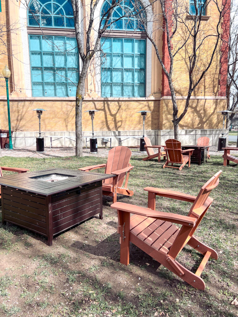 Wooden Adirondack chairs arranged around square fire pit tables with patio heaters outside a historic brick venue, designed by In The Event.